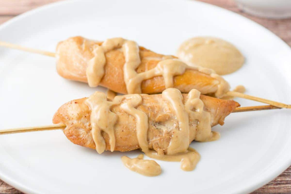 Two chicken tenders on wooden skewers with peanut sauce drizzled on them