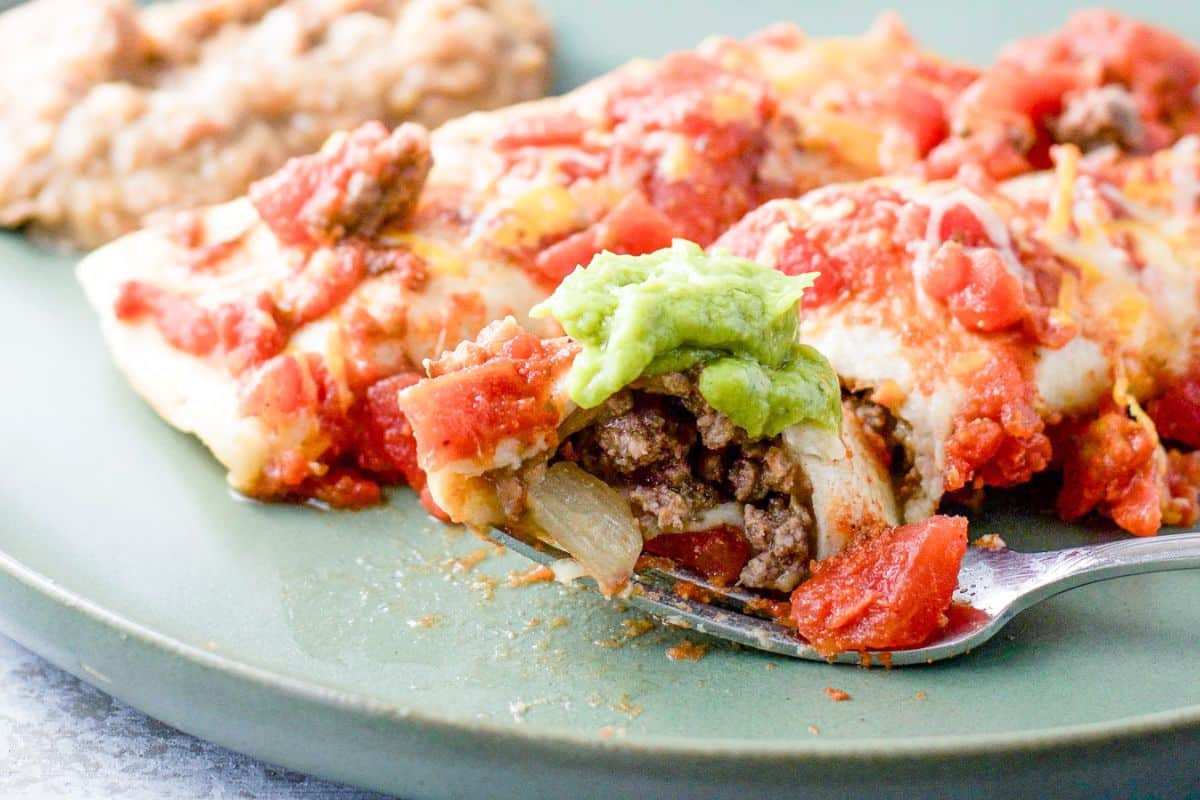 A fork with a bite of enchilada resting on a plate with two enchiladas and beans. There is guac and more enchiladas in the background