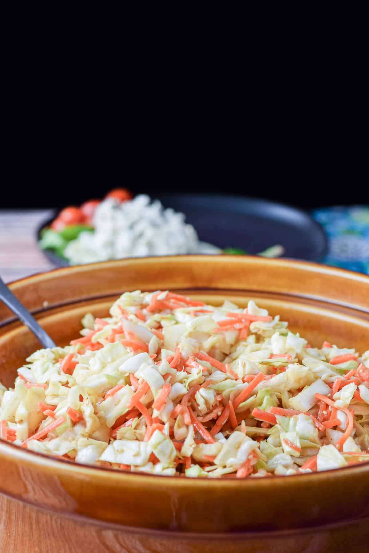 coleslaw mixed in a big brown bowl with a black plate with some lunch on it in the background