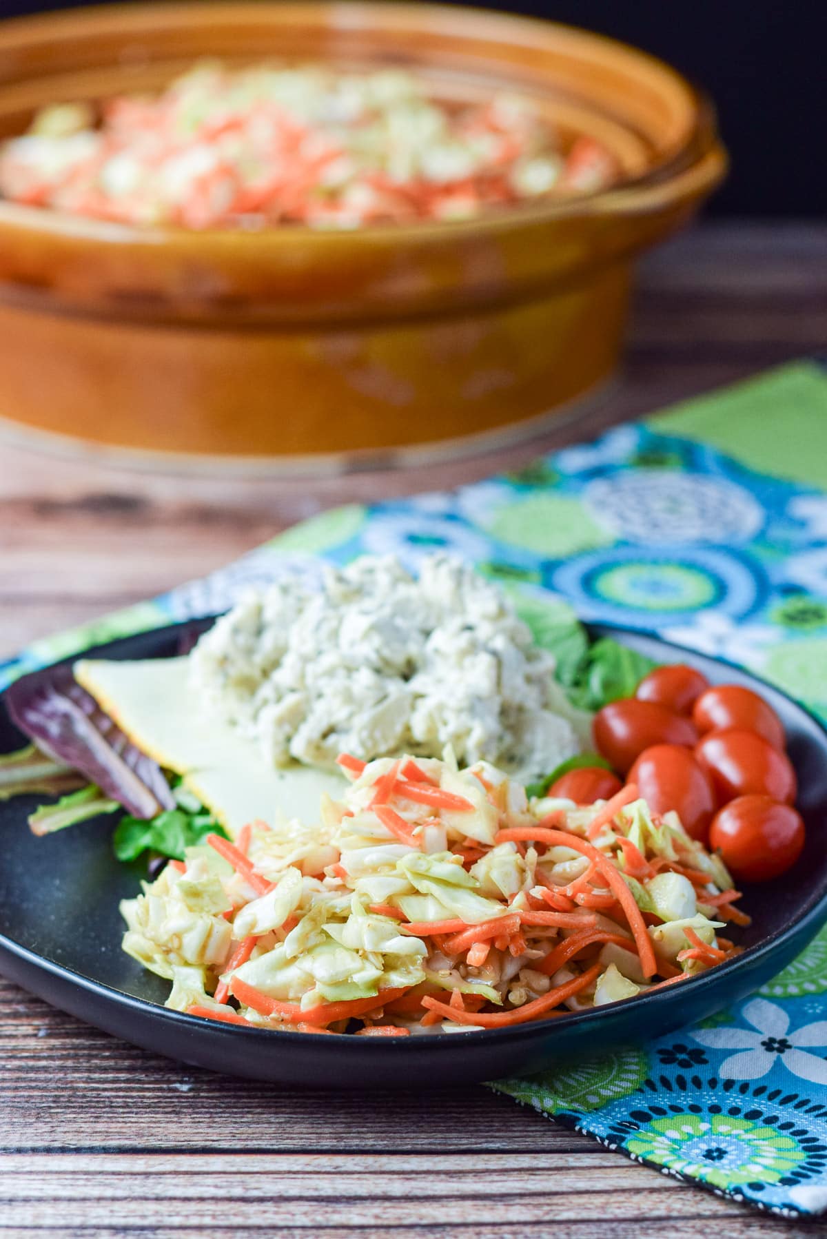 A black plate with coleslaw, chicken salad, munster cheese, lettuce and some grape tomatoes with the big brown bowl of slaw in the background