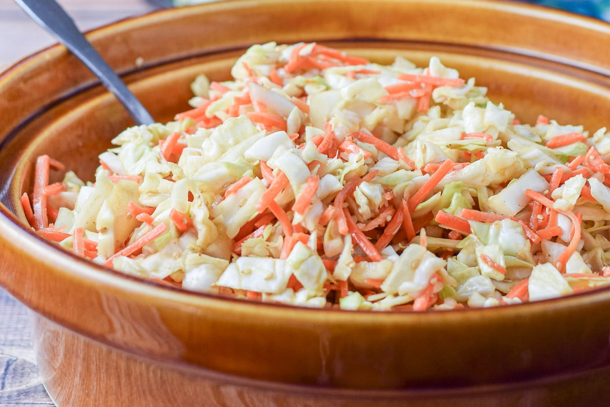 coleslaw mixed in a big brown bowl with a black plate with some lunch on it in the background