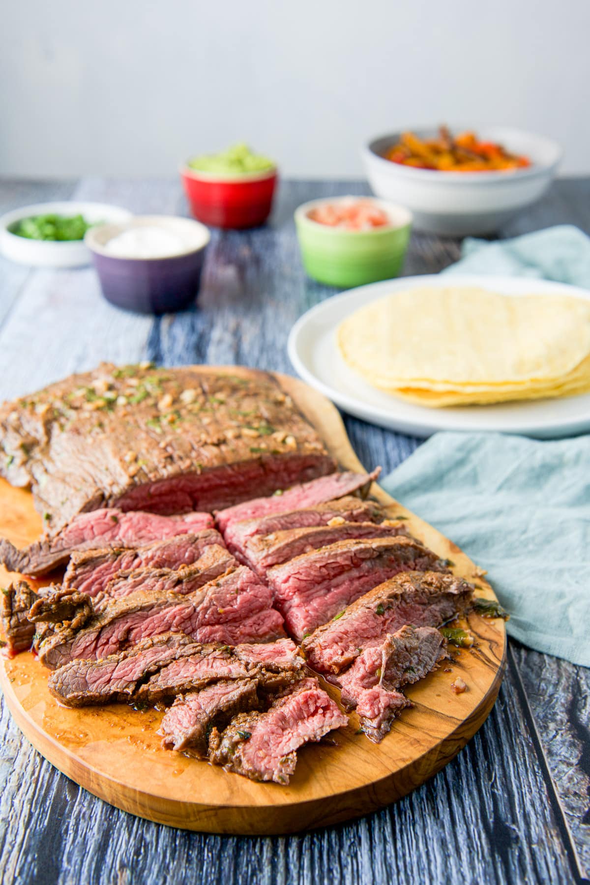 A wooden board with sliced steak, corn tacos, sour cream, cilantro, guac, and veggies