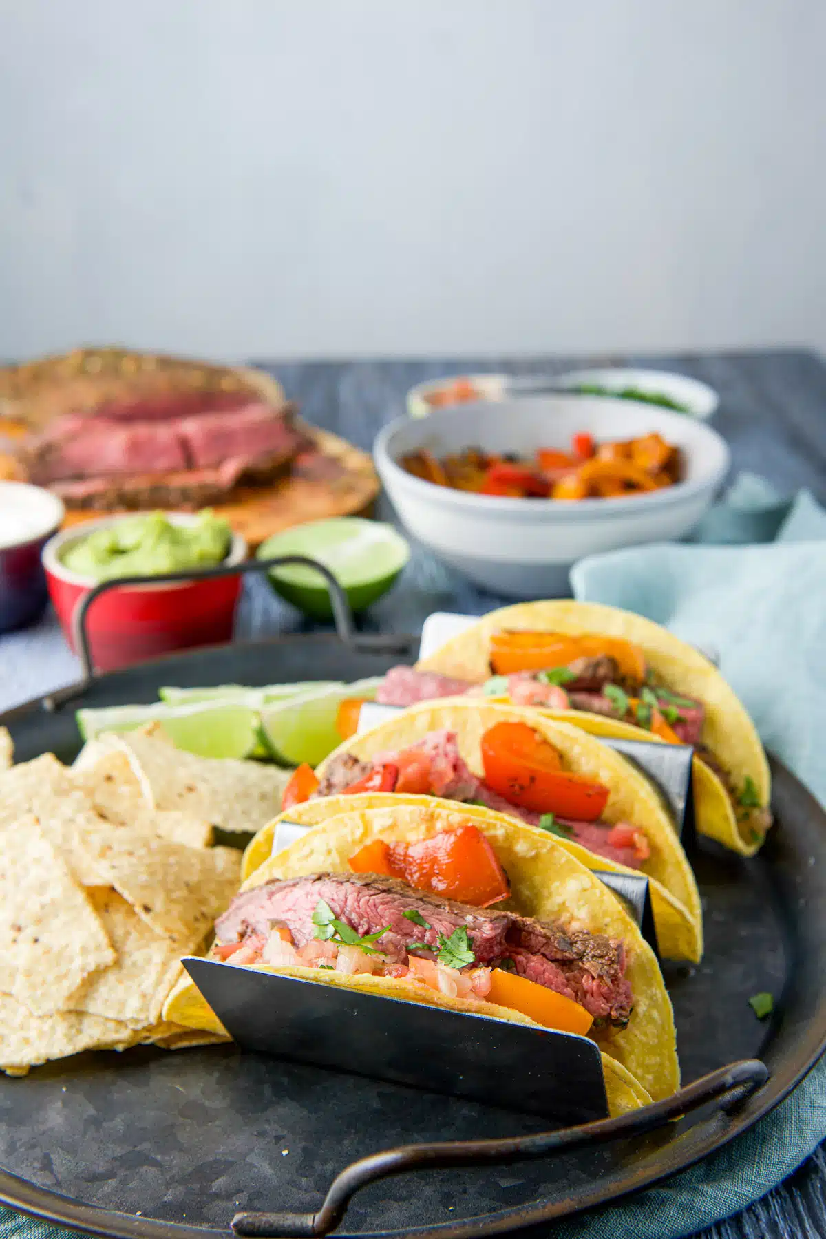 A wooden table with a tray of tacos, chips and lime wedges along with the meat and other garnishes in the back