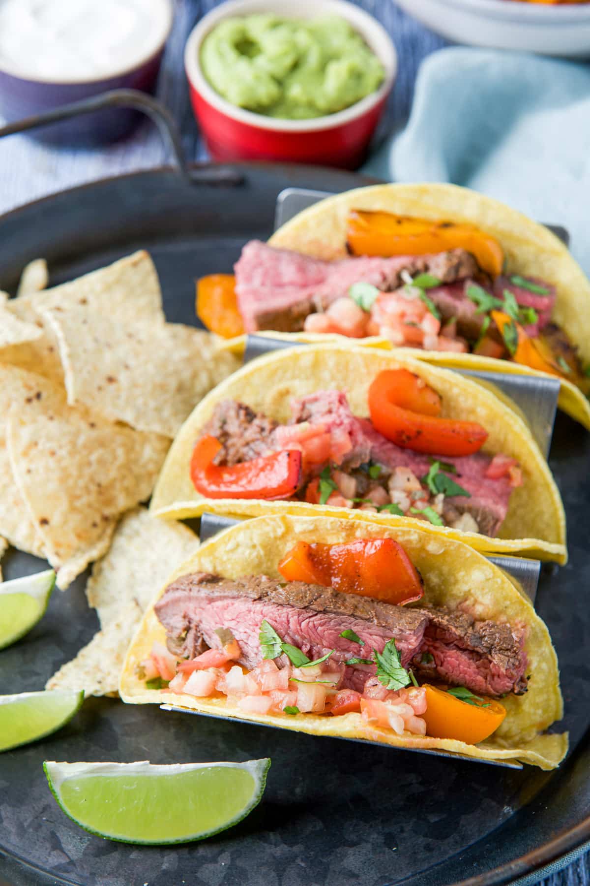Close view of a metal tray with lime wedges, chips and steak tacos with guac and sour cream in the back