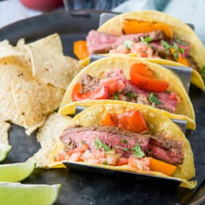 A square photo of a metal tray with three steak tacos on it along with chips and lime wedges