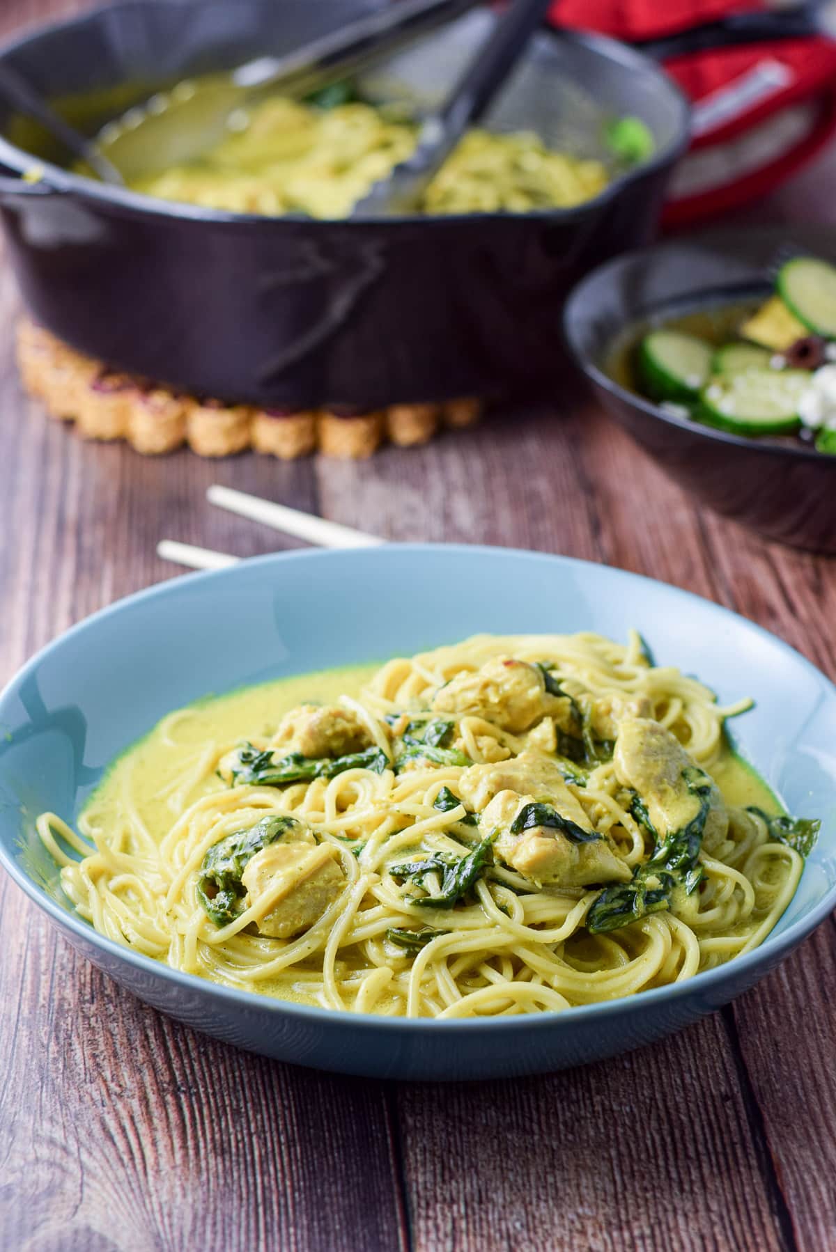 A blue deep dish with noodles, spinach, chicken, and curry sauce - what a pan of the meal behind it