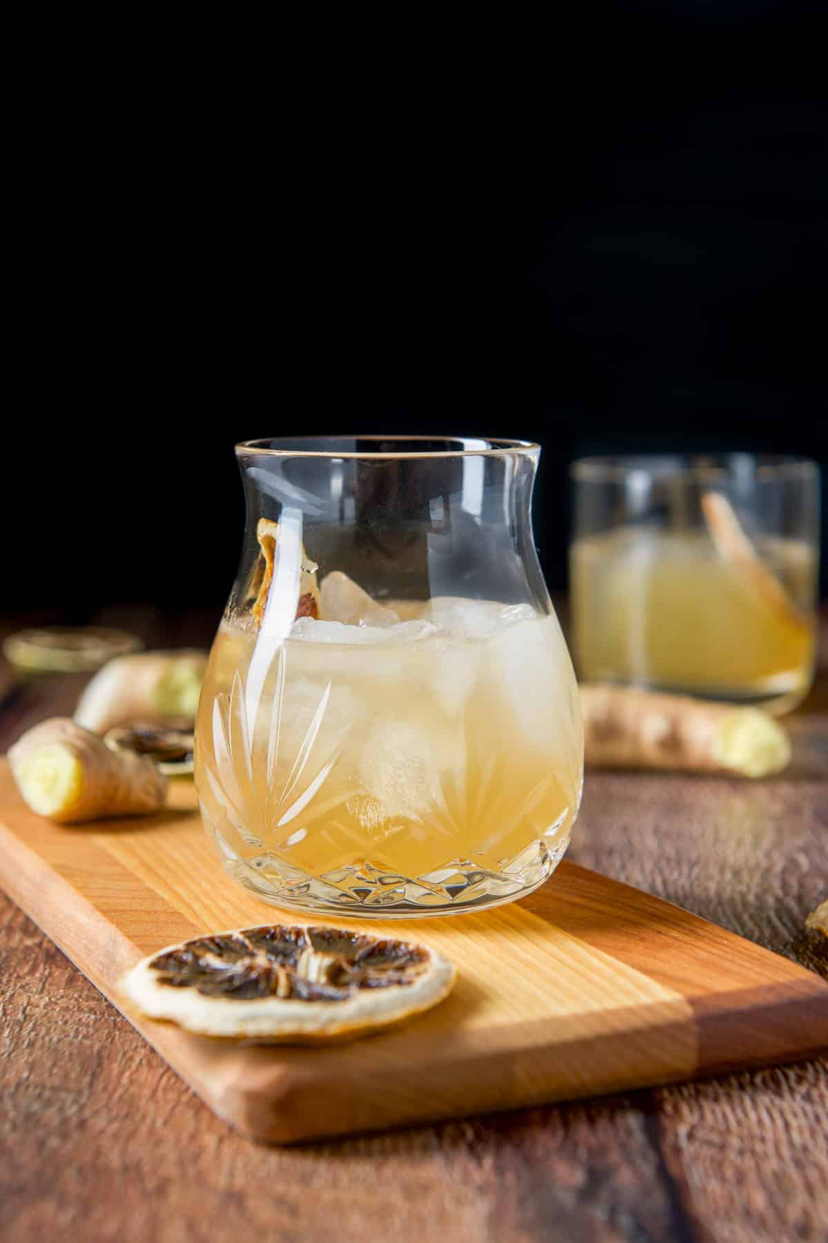 Two vertical views of the amber cocktail in glasses with ginger on the table along with lemon wheels
