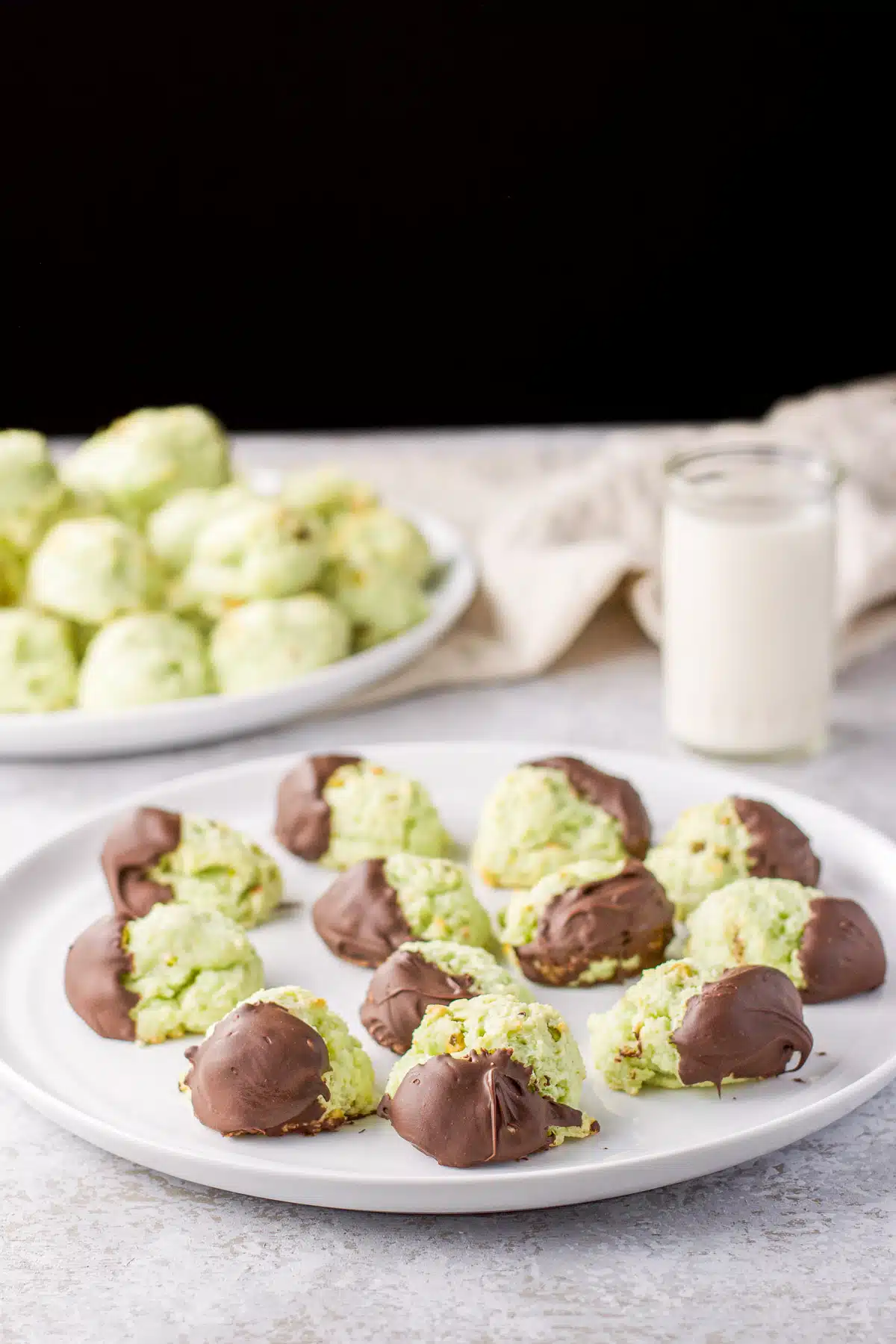 Chocolate dipped pistachio cookies on a plate with other in the back not dipped