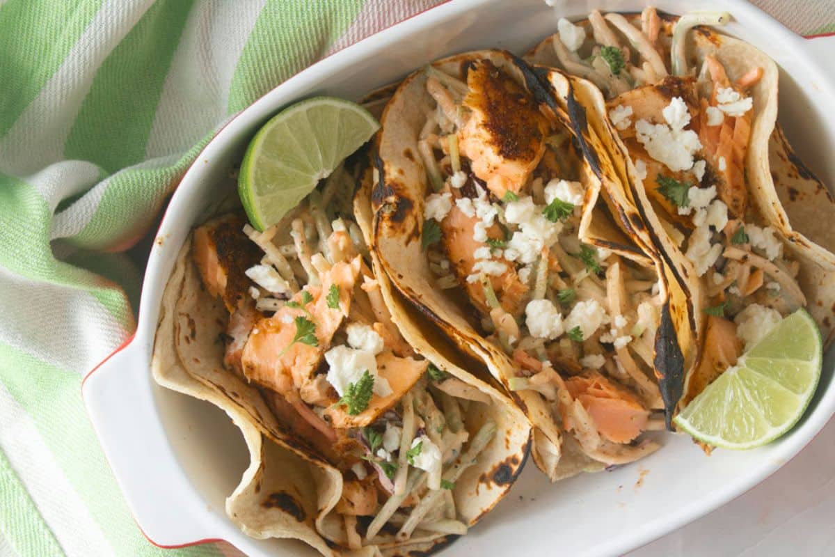 A plate with three salmon tacos and two slices of limes on the sides.