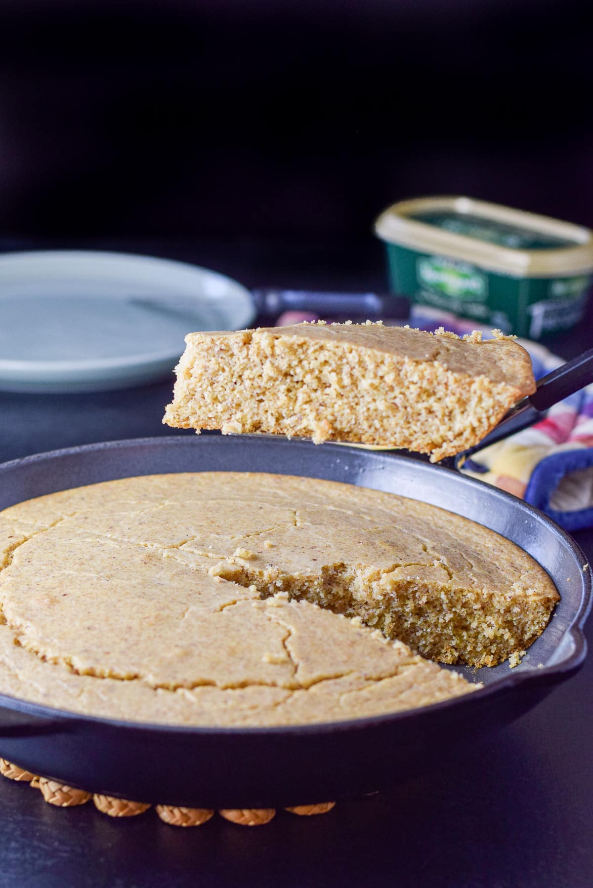 A spatula holding a slice of cornbread over the pan with a plate and butter in the background