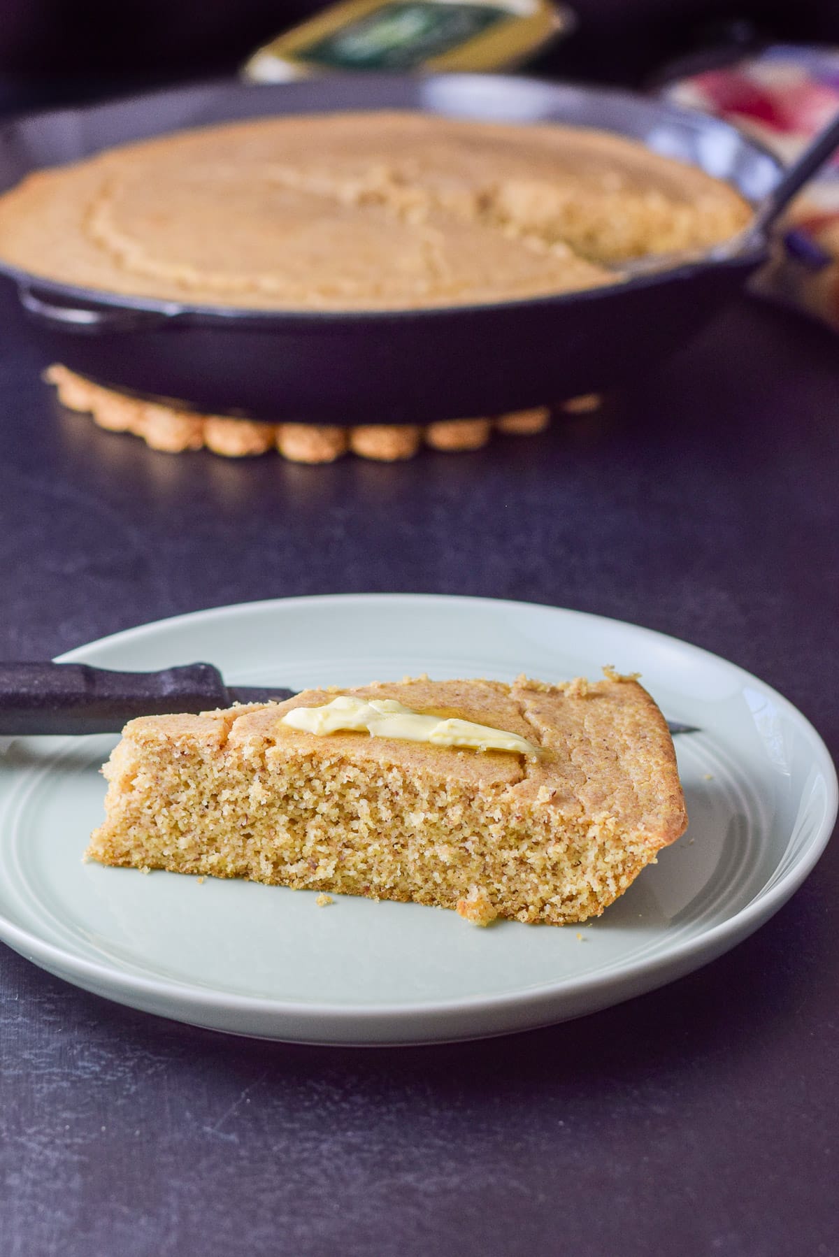 A plate with cornbread on it along with a knife and some butter melting. The pan of cornbread is in the background
