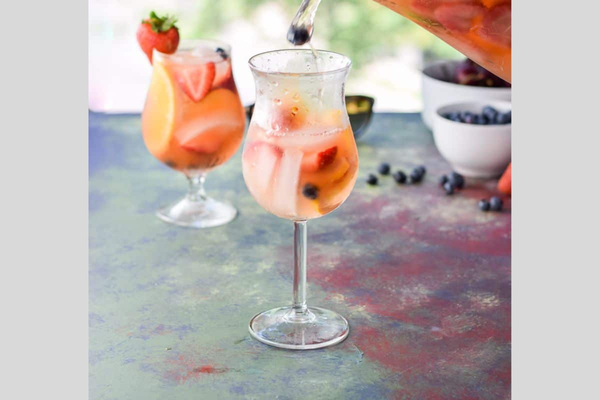 Two glasses on a colorful board with sangria in them. The front glass has a blueberry falling into the glass