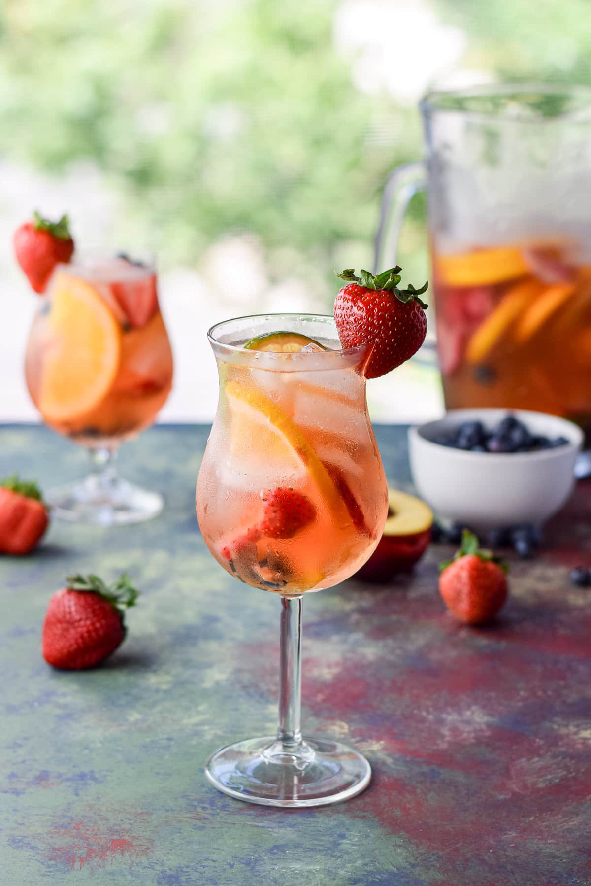 Strawberries on the side of the glasses of sangria with fruit and a pitcher in the background