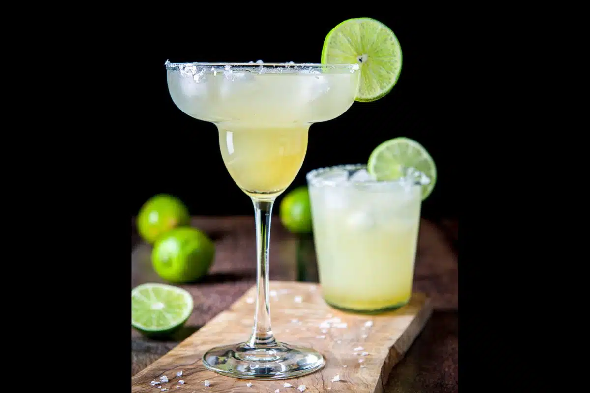 two margarita glasses on a board with salt sprinkled around. Both have a golden margarita in them with lime as garnish