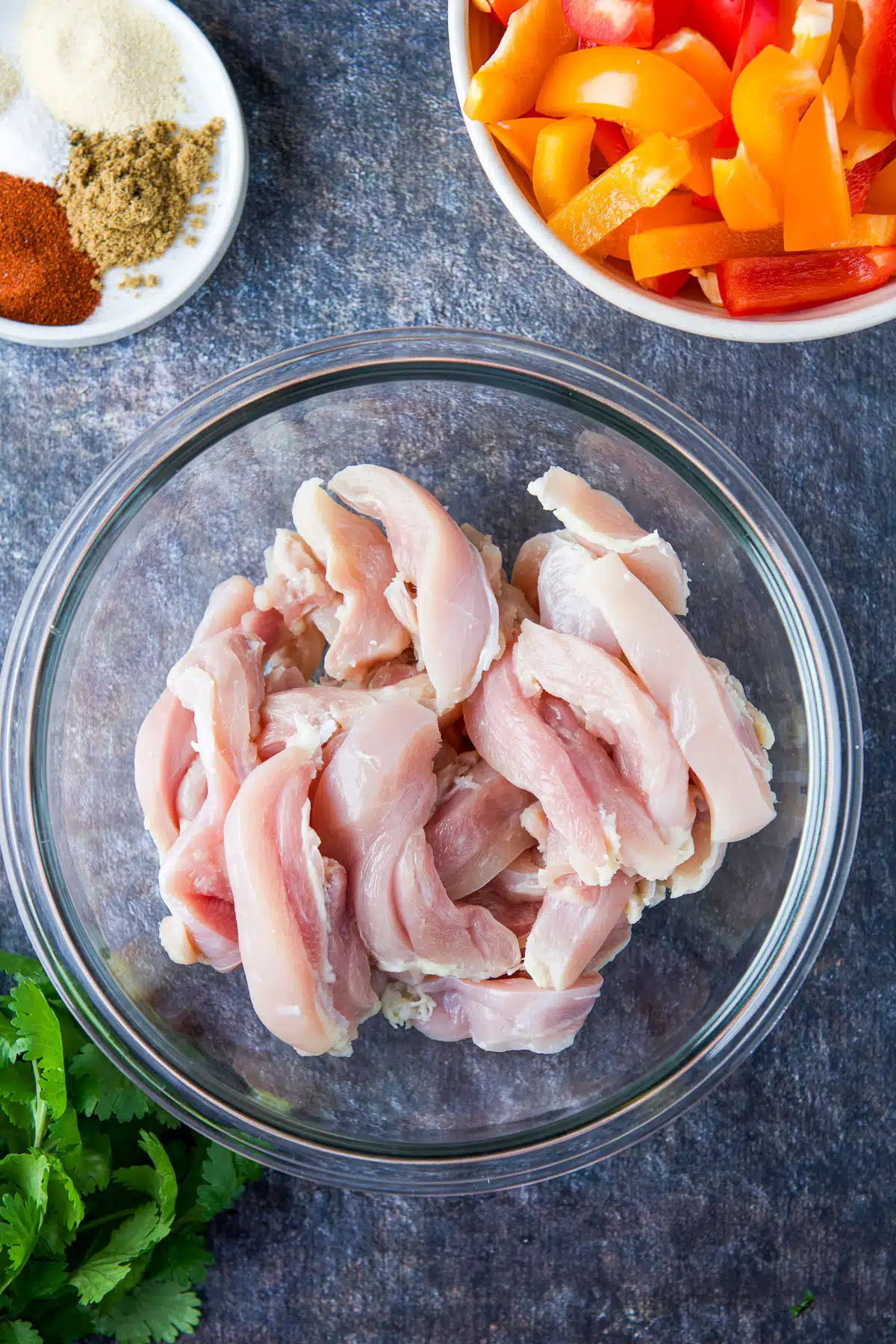 overhead view of raw chicken in a glass bowl herbs and spices, and cut up bell peppers
