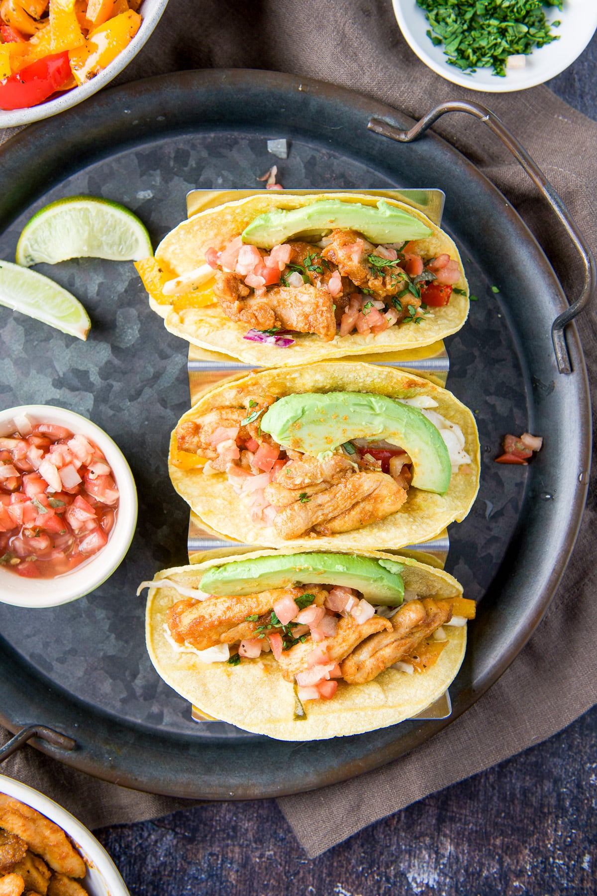 overhead view of the chicken tacos on a metal tray with lime wedges, pico de gallo, bell peppers, and cilantro