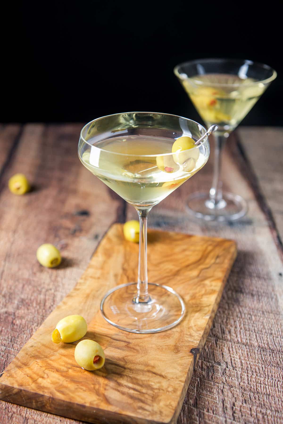 A board with olives on them along with a curved martini glass with another in the back