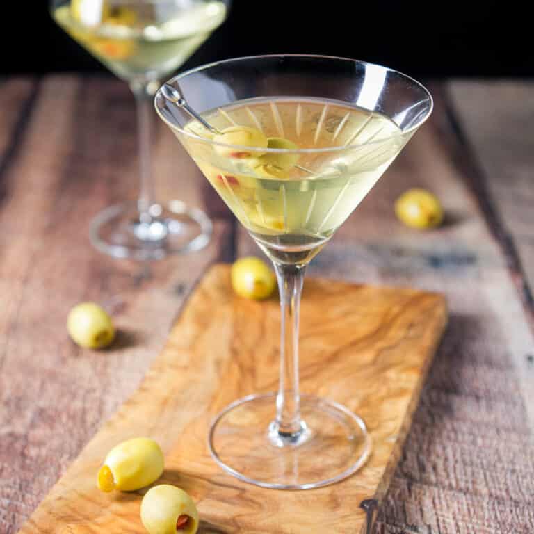 square photo of A beveled martini glass on a small board with another glass in the back, with olives as garnish
