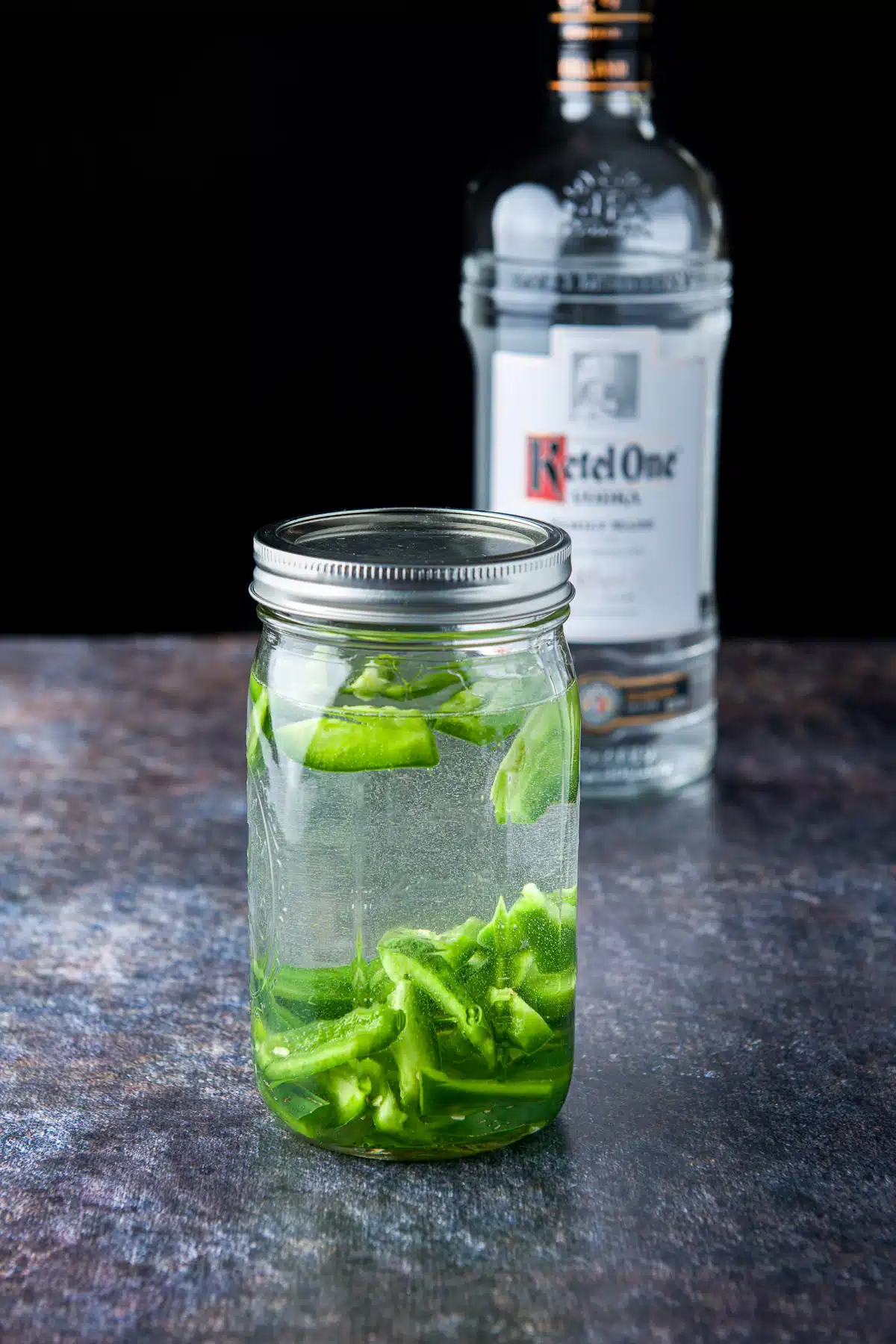 A sealed jar with shaken vodka and jalapeno slices along with the bottle of vodka in the back
