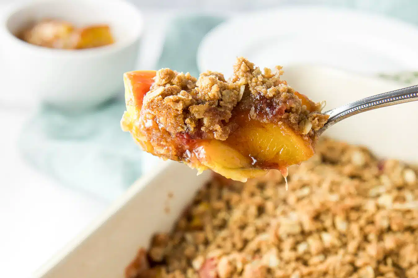 A spoon with peach crisp on it over the pan of crisp with a bowl of it in the back