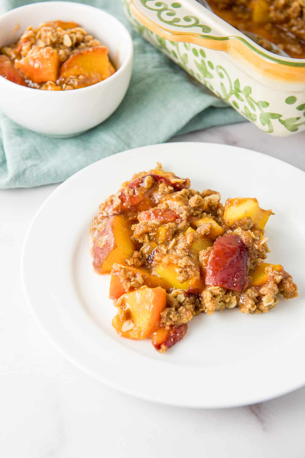 Close up of the peaches mixed with a crumb topping on a plate with a bowl and baking dish in the background