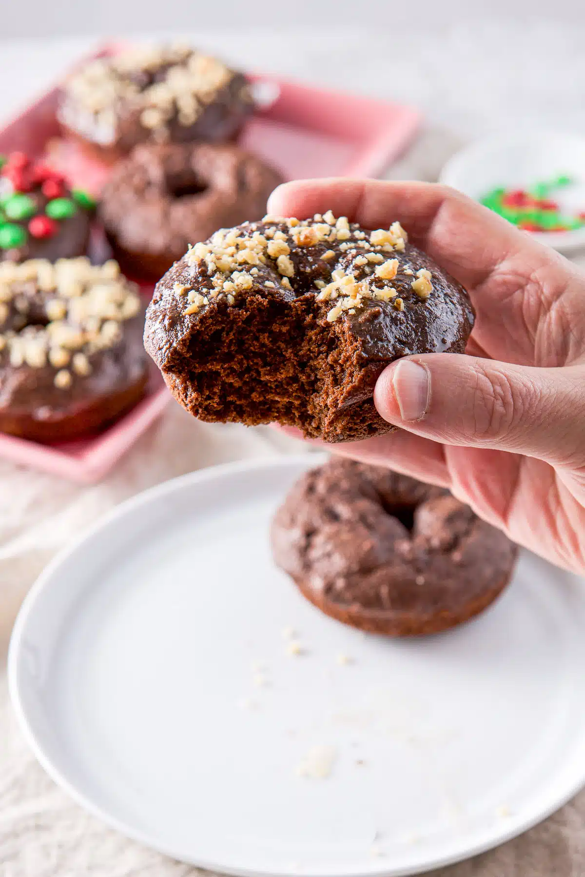 A male hand holding a chocolate donut with walnuts on it. It's held over a white plate and there is a pink platter in the back
