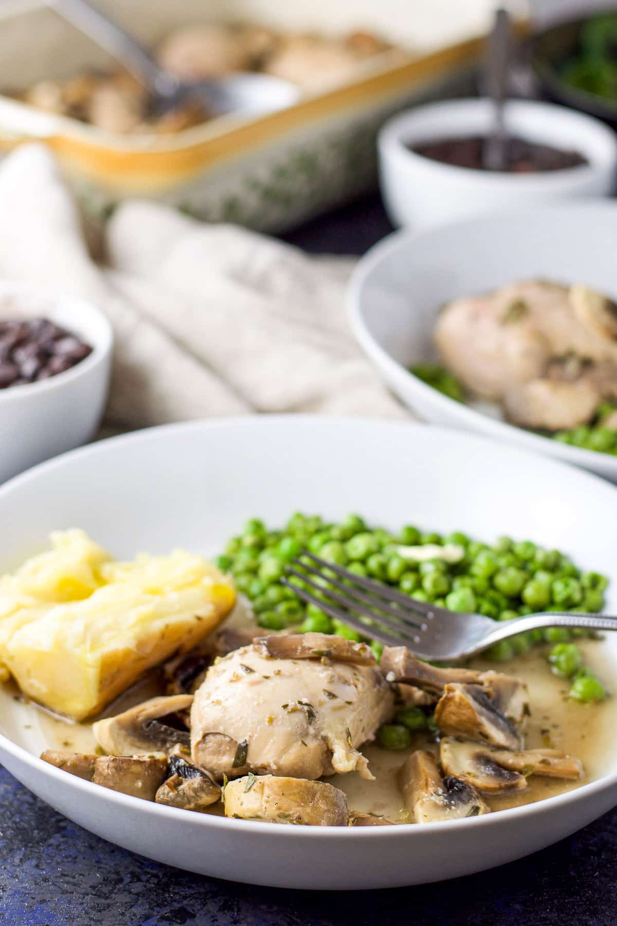 A white deep plate with the chicken, sauce and mushrooms with another plate in the background as well as the baking dish