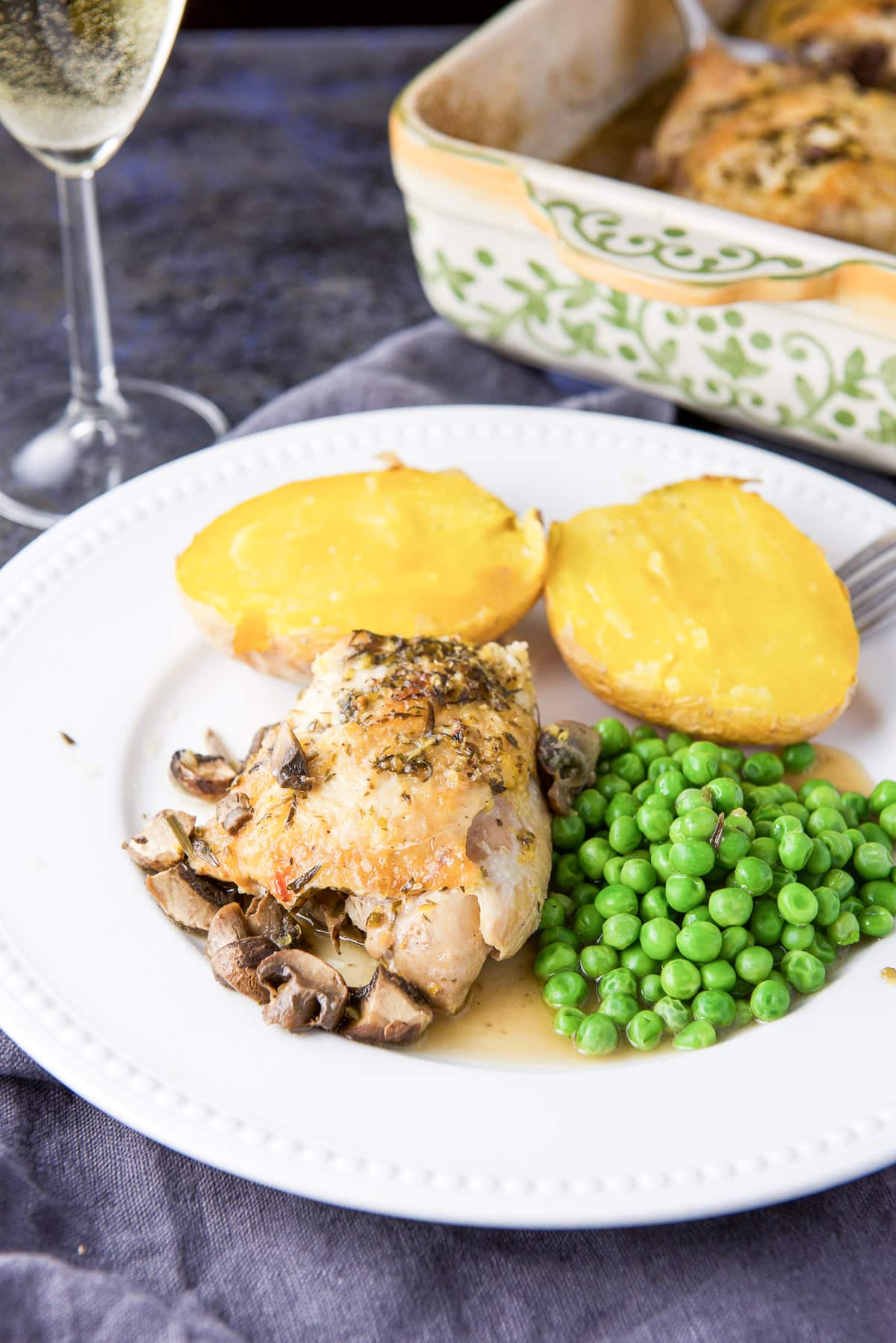 A chicken thigh on a white plate with the baking dish and wine in the background