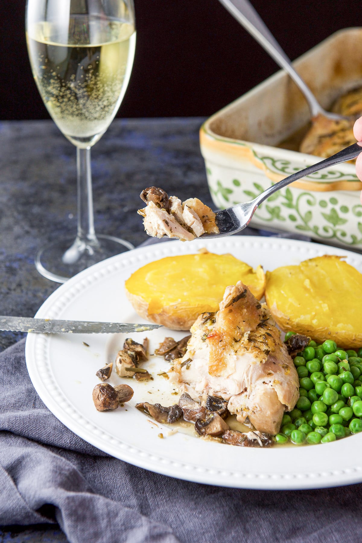 A fork with chicken on it held over the plate with wine and the baking dish held in the background