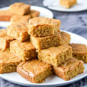 A white plate with a pile of blond brownies on it with others in the back - square