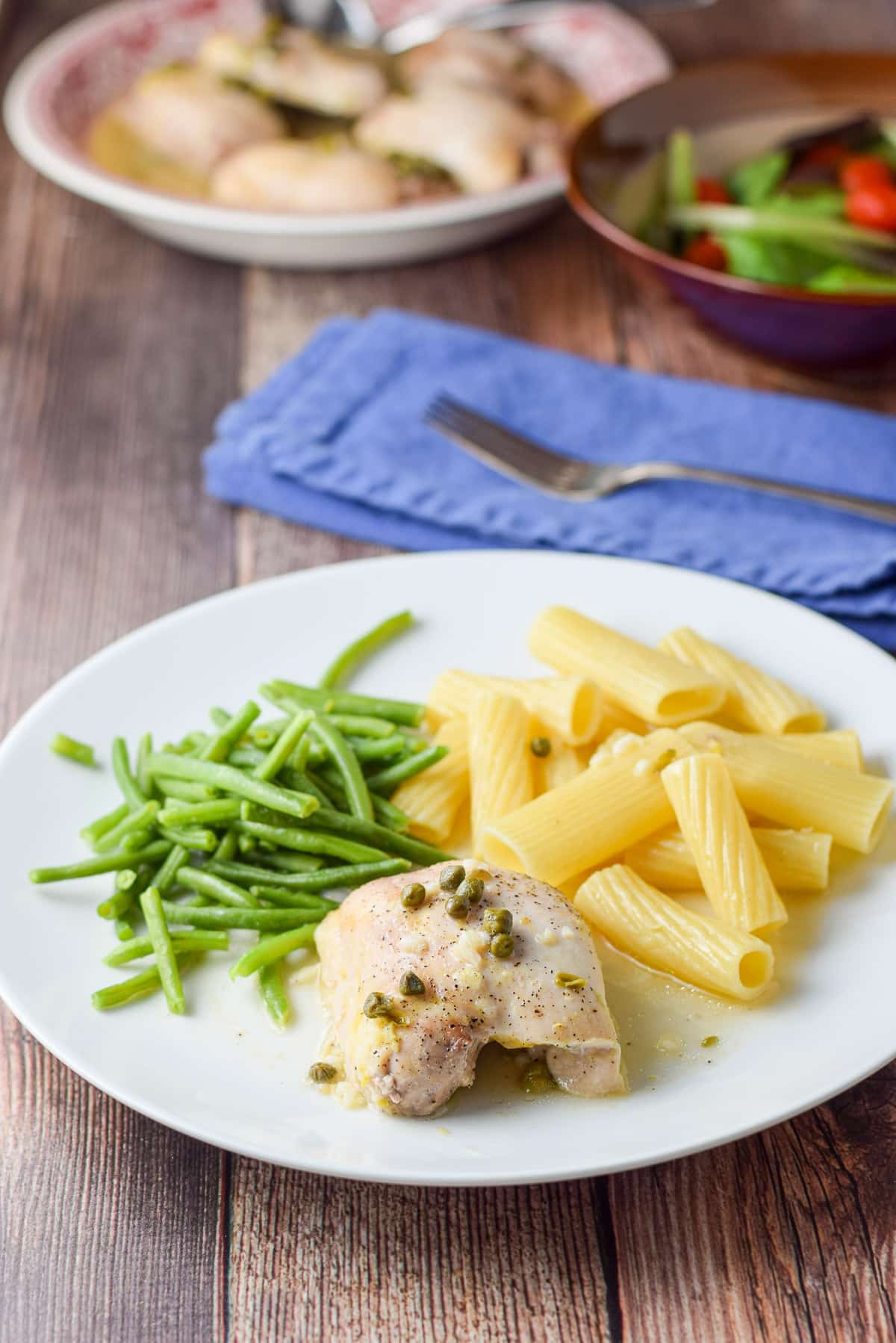A white plate with the piccata, capers, pasta and green beans. There is a salad and a bowl of chicken in the background