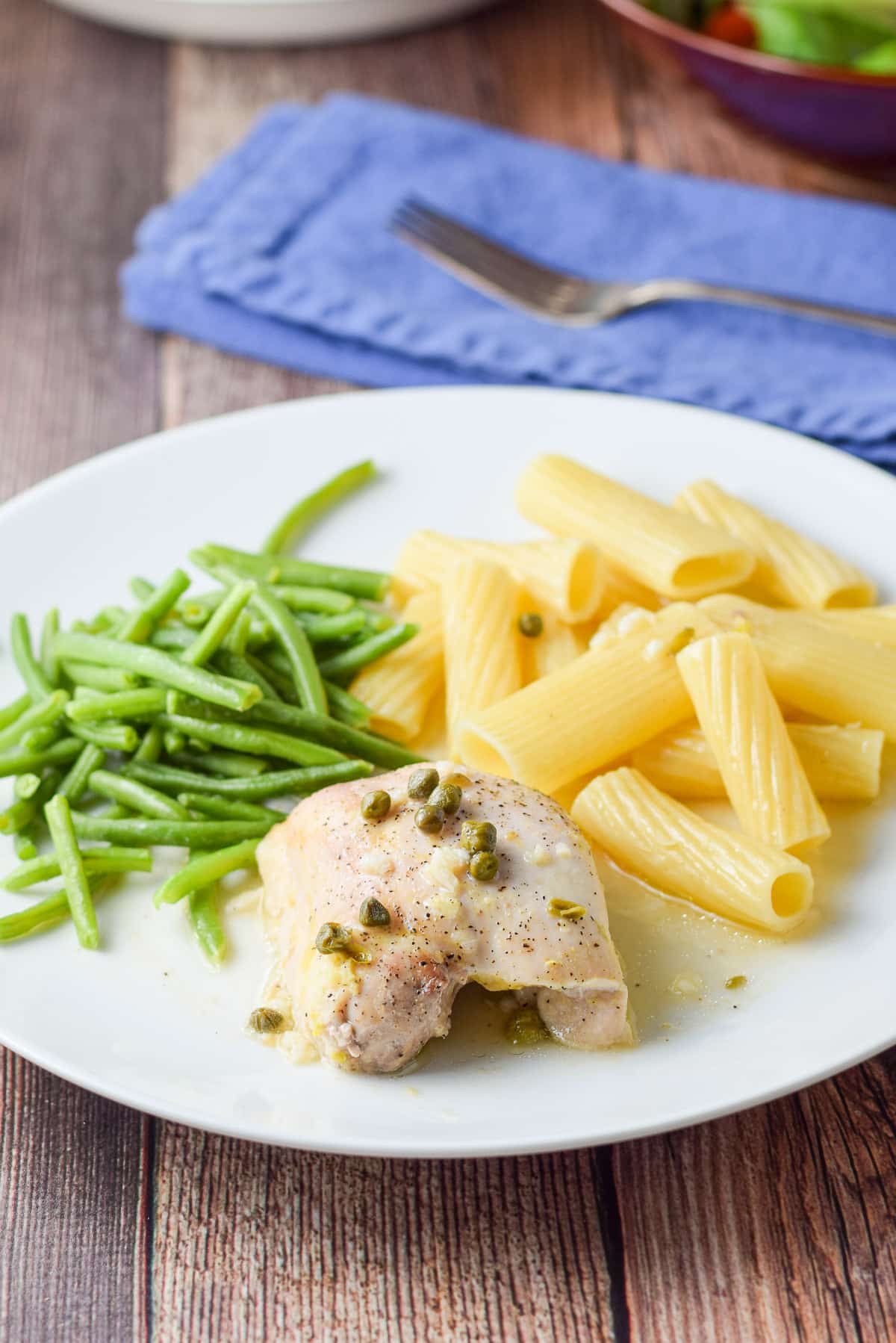 Closeup of a white plate with a chicken thigh with capers on it along with pasta and string beans