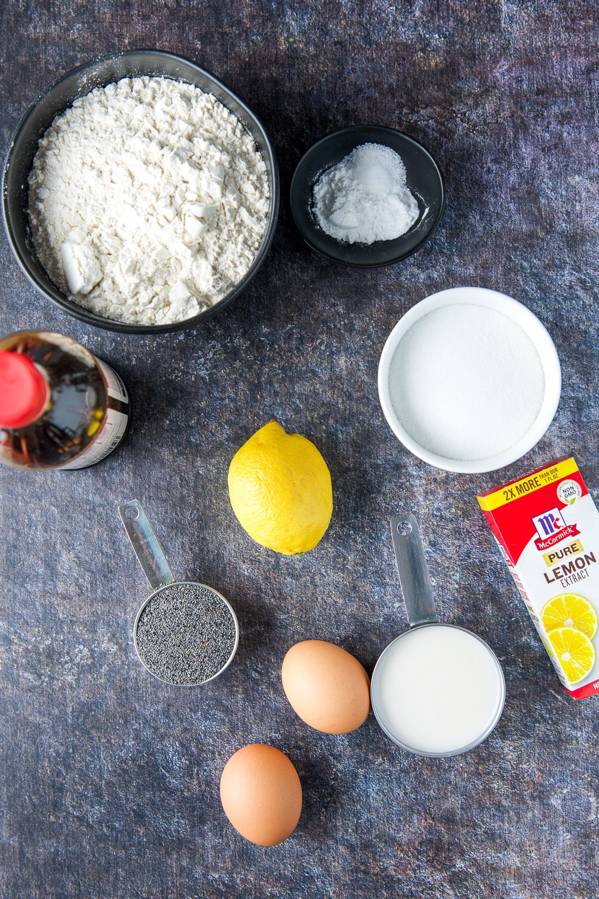 Overhead view of the ingredients - flour, baking soda, baking powder, milk, lemon extract, lemon, eggs, vanilla, and poppy seeds