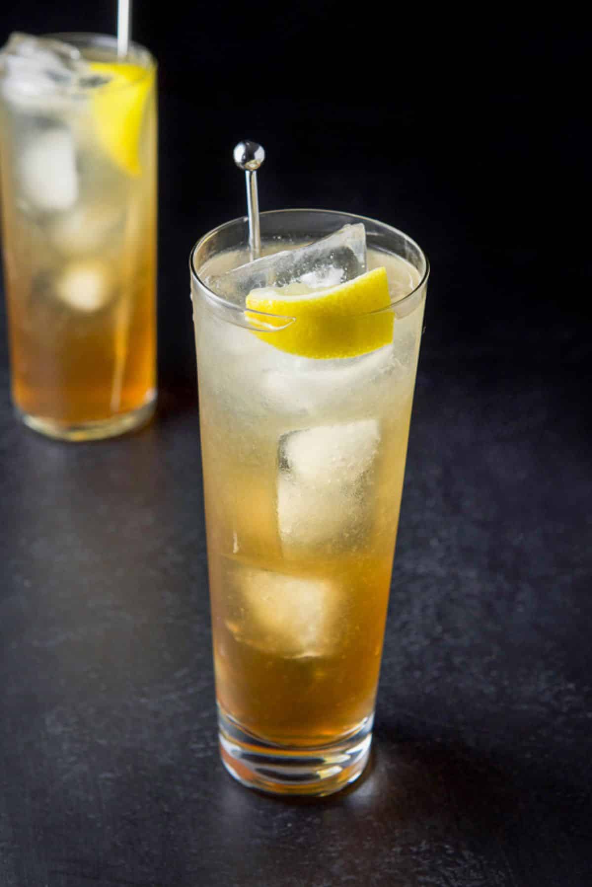 Wider glass in front filled with the cocktail in front of the collins glass with lemon wedges and stirrers in them