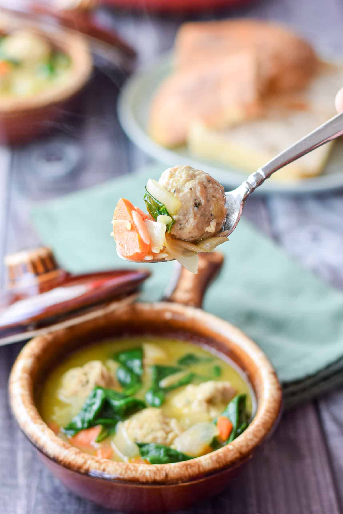 A spoon holding a meatball and vegetables held over a crock of the soup
