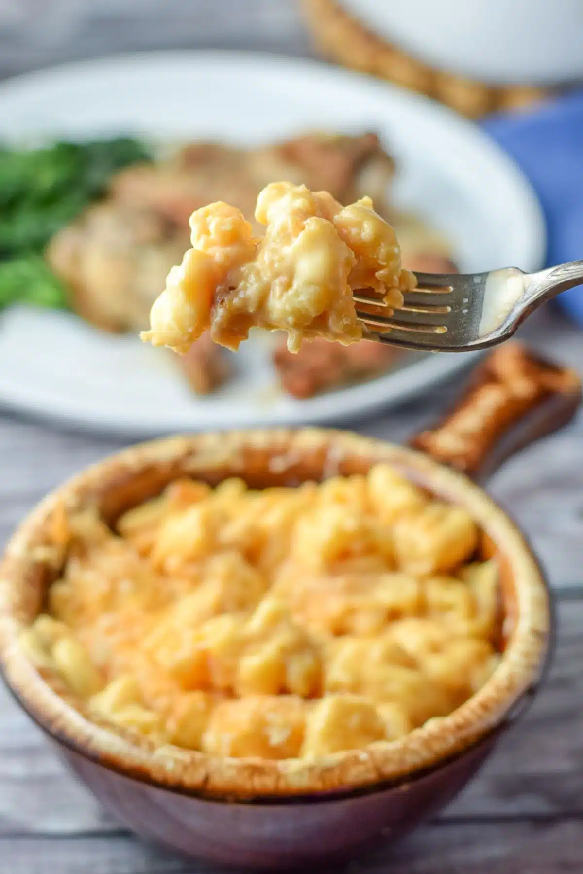 A forkful of cheesy macaroni over a crock filled with it - also with a plate in the background