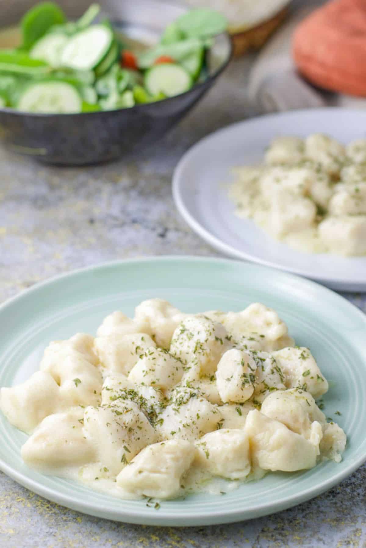 A green round plate with gnocchi with cream sauce on it with another plate, salad and casserole dish in the background