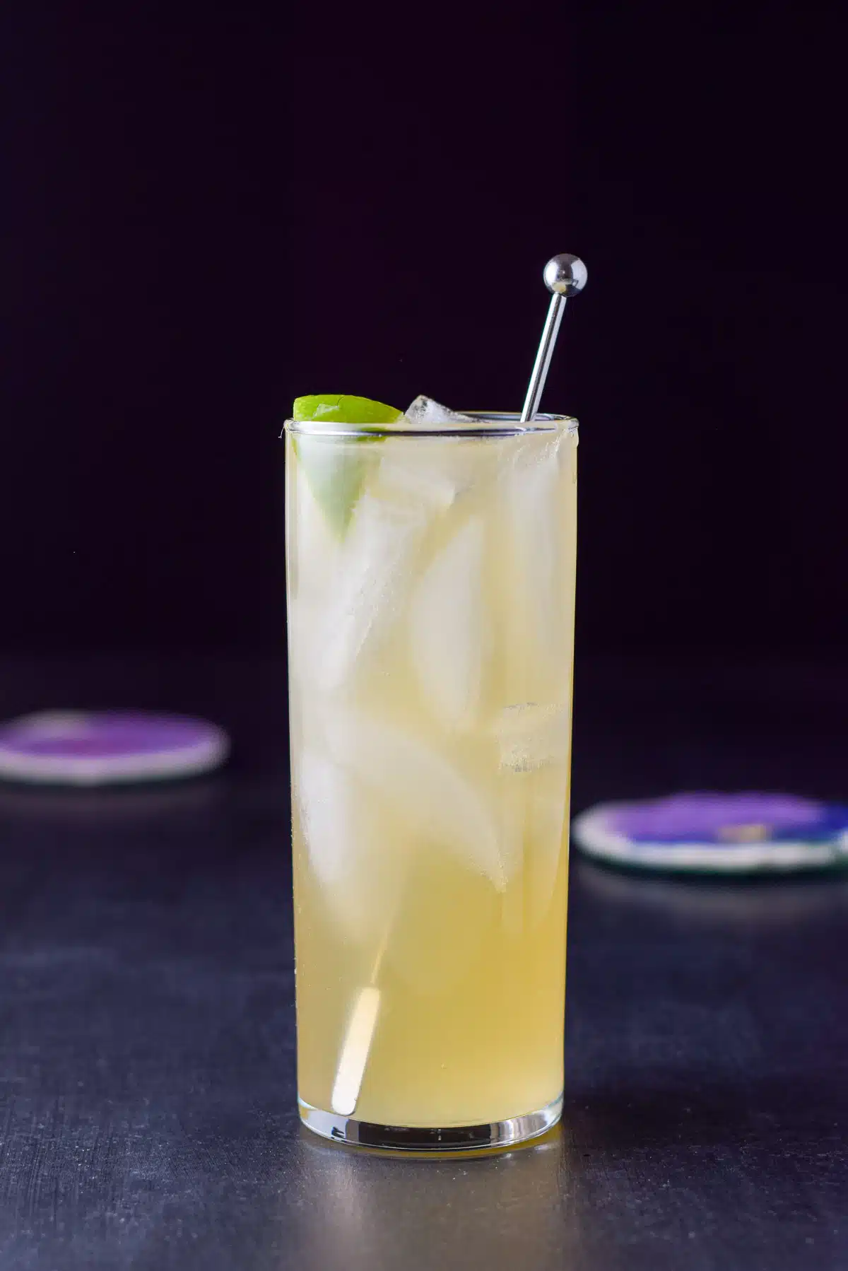 Vertical view of a collin glass with the yellow cocktail with a lime wedge and stirrer in it