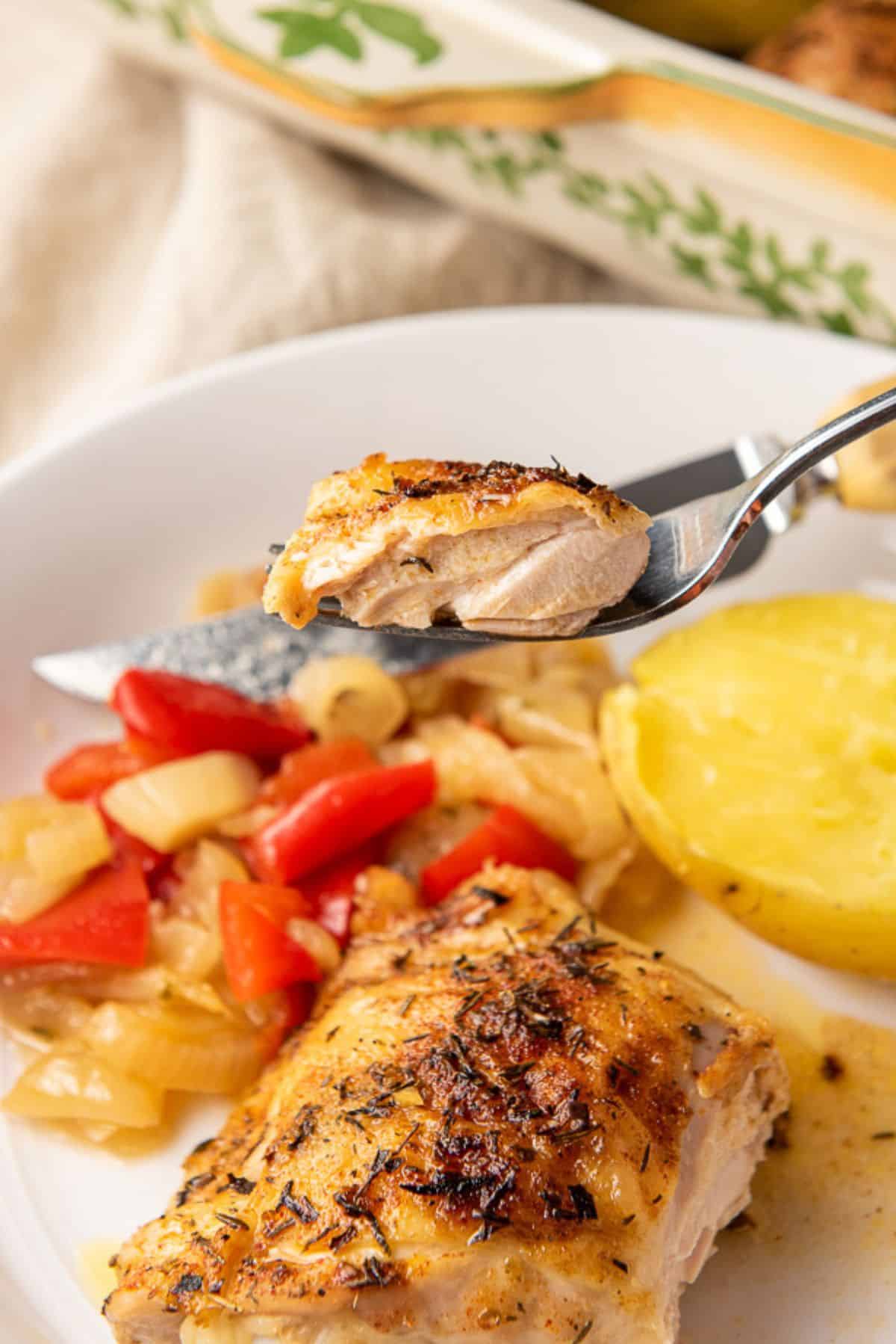 A bite of chicken thigh on a fork held over the dish with onions and potato