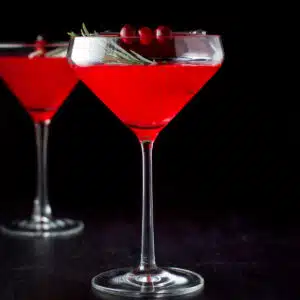 a square photo of two glasses filled with a cranberry cosmo, with cranberries and rosemary for garnish
