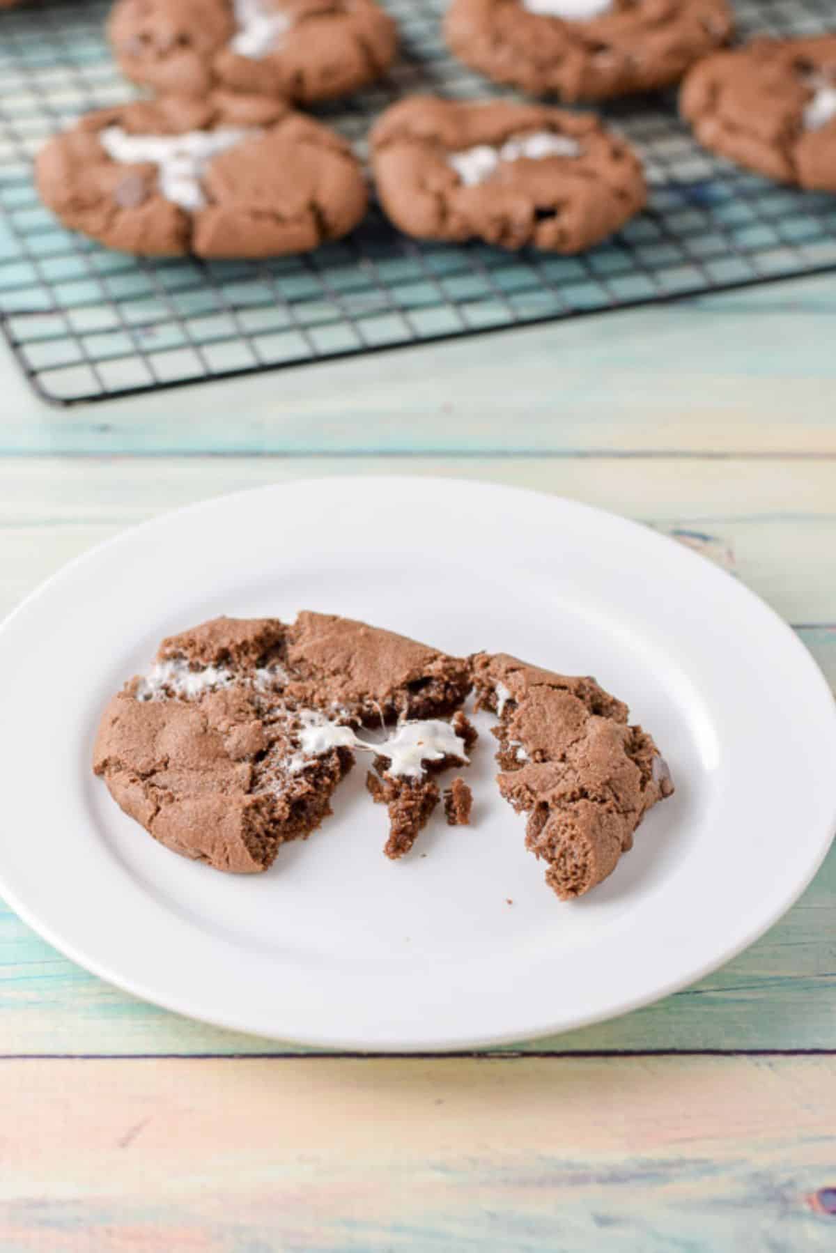 A white plate with a broken up marshmallow chocolate cookie with more cooling in the back