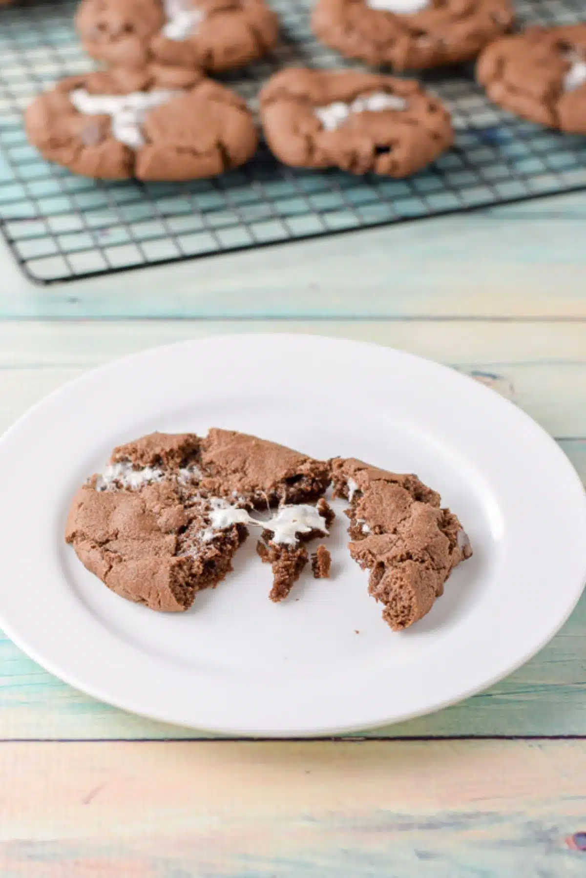 A white plate with a broken up marshmallow chocolate cookie with more cooling in the back