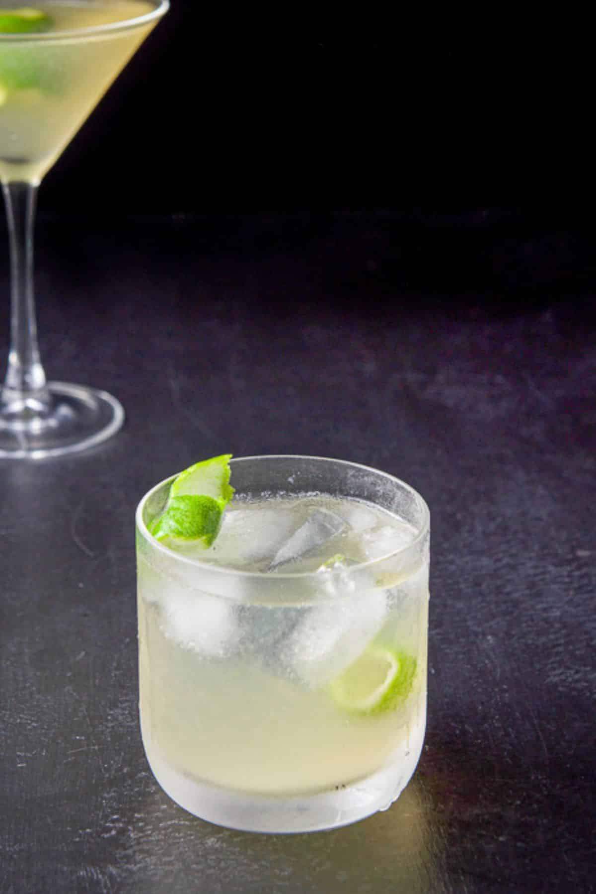 Double old fashioned glass in front of the classic glass filled with the gimlet and lime twists
