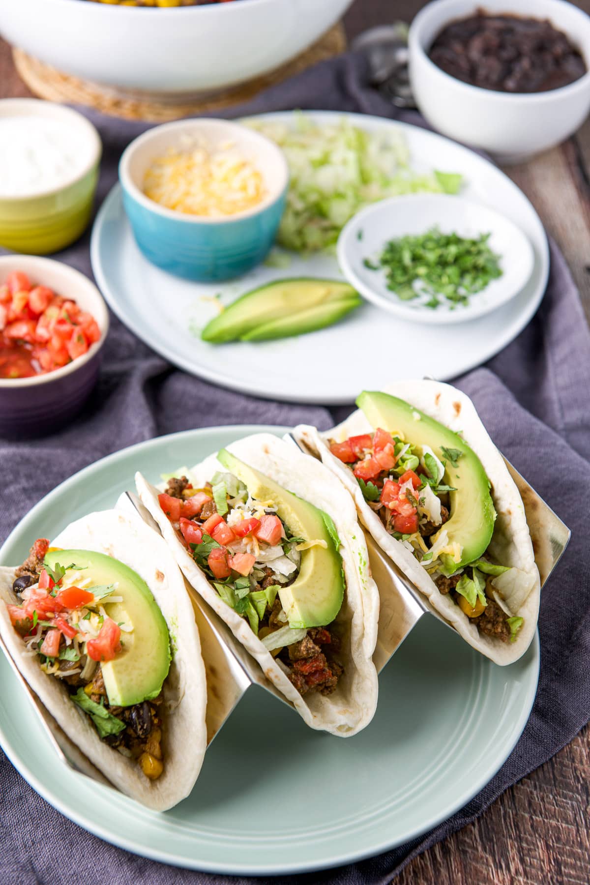 A green plate with a taco holder with three soft tacos in it with all the fixings in the tacos and in the background
