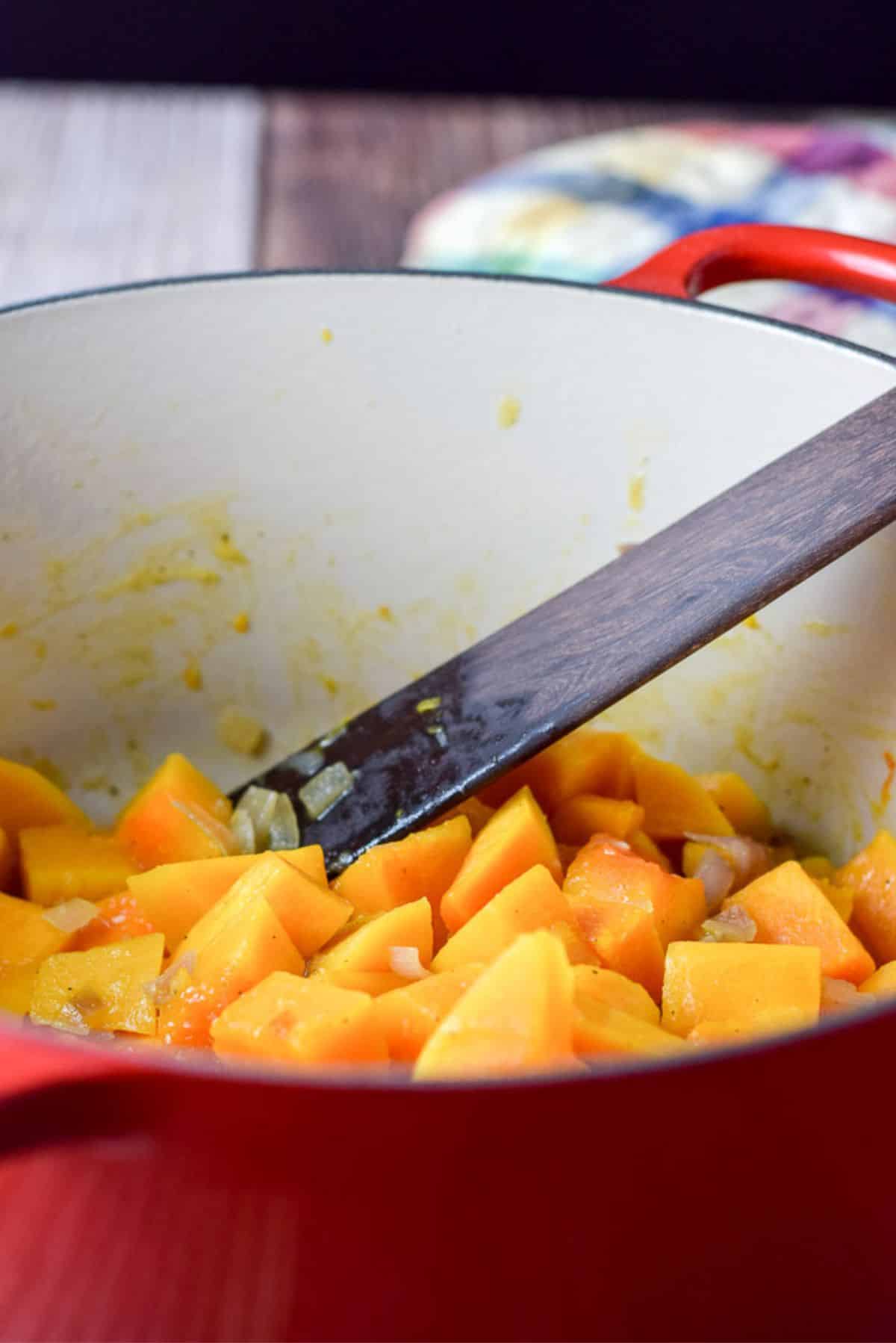 Cut squash cooked in chicken broth with some onions in a red pot and a wooden spoon in the pot