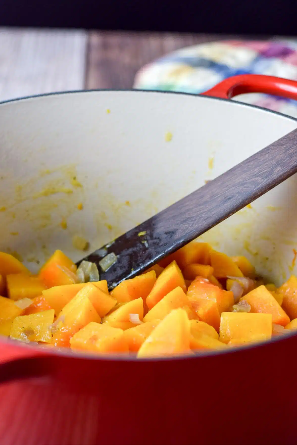 Cut squash cooked in chicken broth with some onions in a red pot and a wooden spoon in the pot