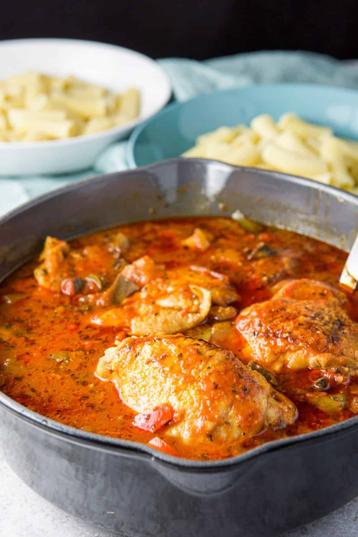 Cast iron pan filled with chicken cacciatore with two bowls of pasta in the background