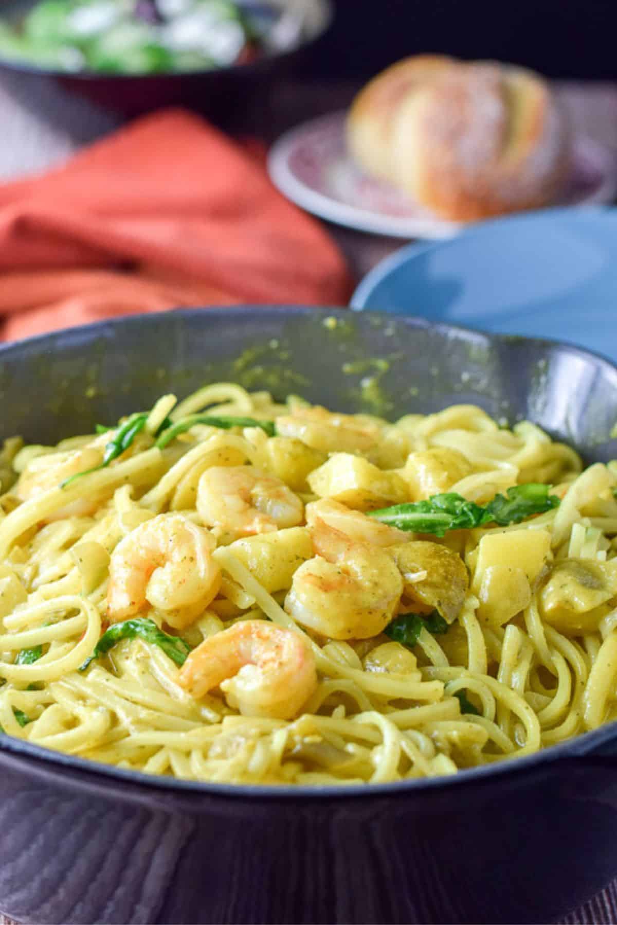 The shrimp on curry noodles in a big pan with a roll, plate and salad in the background