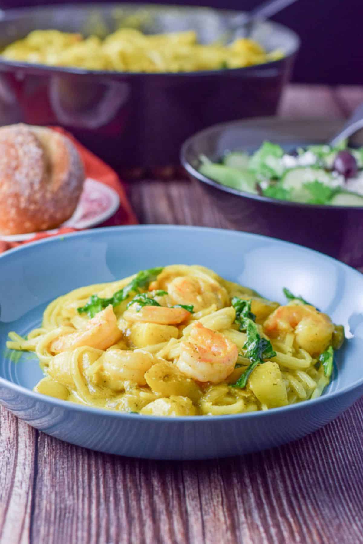 Shrimp with curry sauce and noodles in front of a pan, bread and bowl of salad