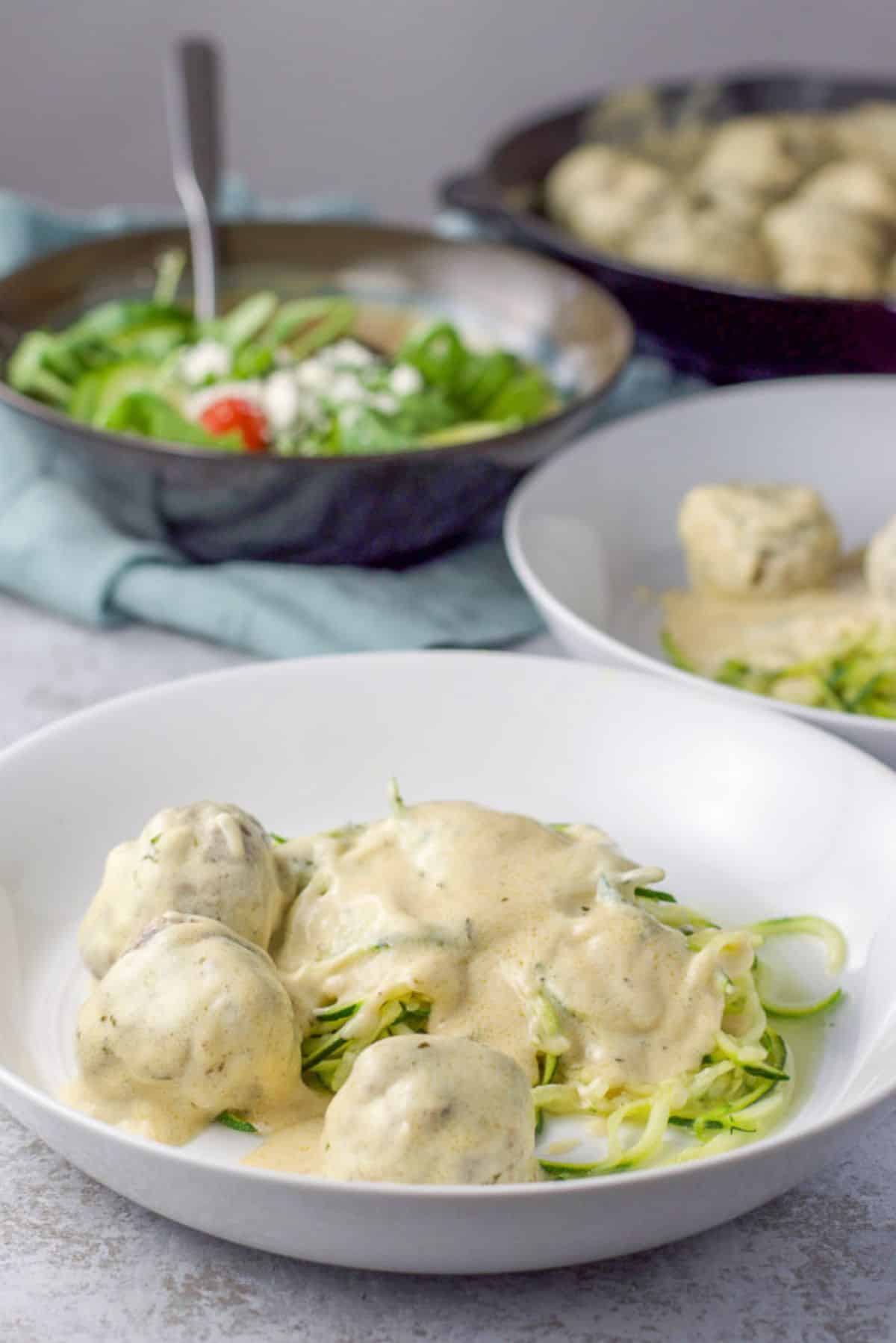 Two deep plates of meatballs with a cream sauce on zoodles.There is also a salad in the background and a pan of the meatballs